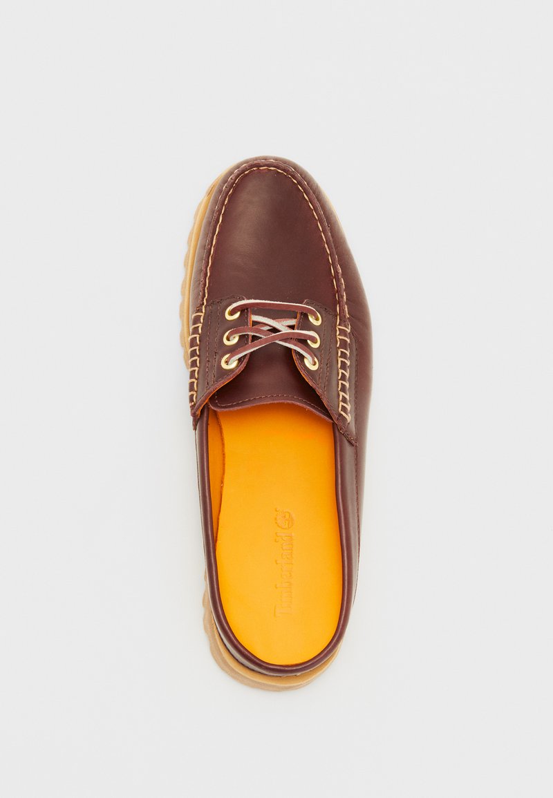 Scarpa slip-on in pelle marrone con suola beige, caratterizzata da occhielli dorati, cuciture crema e soletta arancione. Superficie liscia con punta arrotondata.