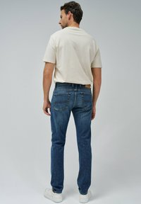 Jeans de mezclilla azules para hombre con corte recto, bolsillos traseros y una camiseta ligera beige, combinados con zapatillas blancas. Diseño sencillo, ajuste casual.