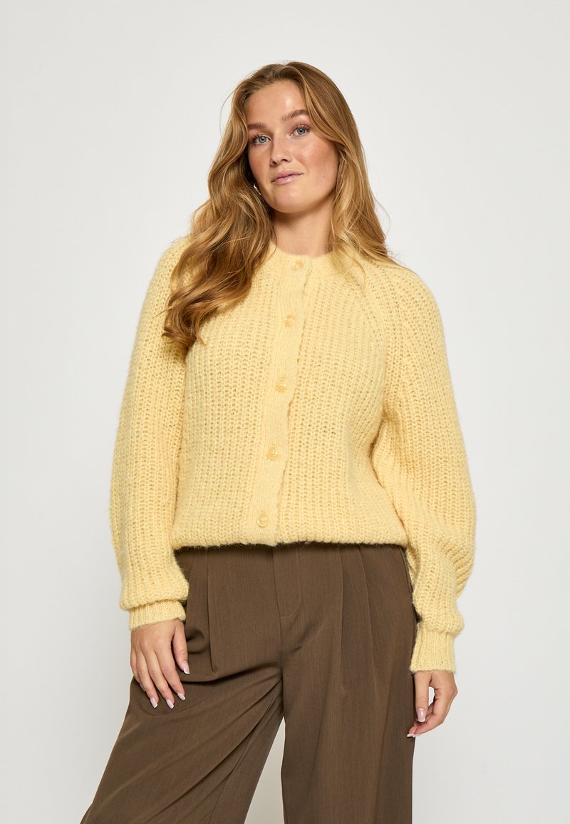 Dzeltena adīta jaka ar kuplu tekstūru, apaļu kaklu un pogu aizdari. Iezīmējas ar garām piedurknēm un ribota akcenta apdari pie rokai un apakšmalas.