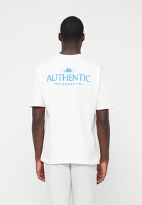 Man die met zijn rug naar de camera staat, gekleed in een wit T-shirt met blauwe tekst en logo "AUTHENTIC" op de achterkant, gecombineerd met lichtgrijze broek.