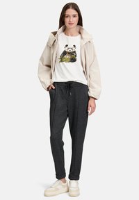 Hellbeige Jacke über einem weißen T-Shirt mit Pandagrafik, kombiniert mit dunkelgrauen Sweatpants und beigen Turnschuhen. Lässiges urbanes Outfit.