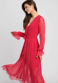 Czerwona sukienka w groszki z długimi, falbankowymi rękawami. Lekki, przezroczysty materiał, z głębokim dekoltem w kształcie litery V i detalem w postaci wiązania w pasie. Luźny, swobodny krój.