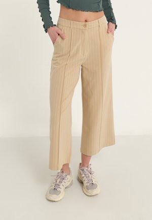 Person wearing beige pinstriped cropped trousers, white and gray chunky sneakers, and a dark green long-sleeve cropped top with ruffled edges.