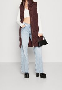 Burgundský péřový vest s vysokým límcem, zipem vpředu a prošívaným vzorem, nošený přes bílý crop top a světle modré široké džíny.