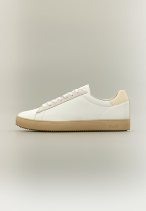 Baskets basses blanches avec accent beige au talon, lacets blancs et semelle en caoutchouc gomme, présentées sur un fond neutre.