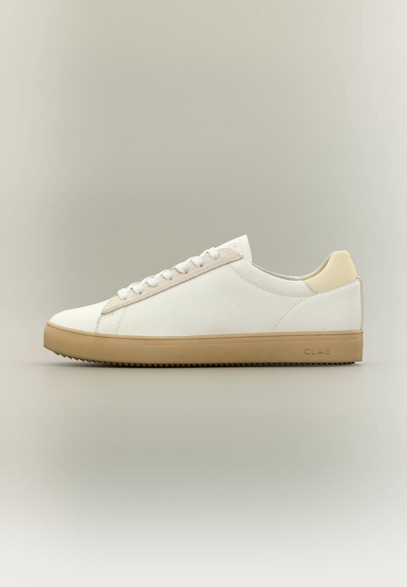 Λευκό sneaker χαμηλού ύψους με μπεζ λεπτομέρεια στη φτέρνα, λευκά κορδόνια και σόλα από καουτσούκ καραμέλας, εμφανίζεται μπροστά σε ουδέτερο φόντο.