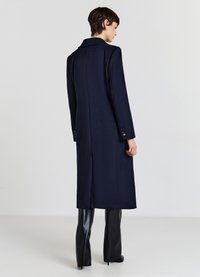 Cappotto lungo in lana blu navy con colletto sagomato, apertura posteriore singola e dettagli a quattro bottoni sui polsini, abbinato a pantaloni neri aderenti.