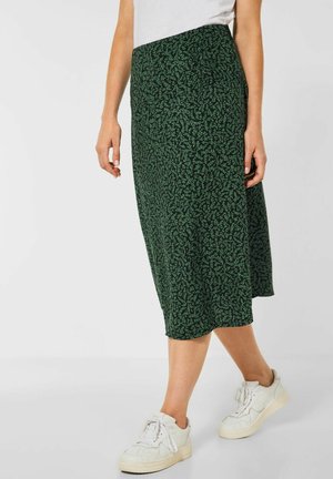Femme portant une jupe midi vert foncé avec un petit motif feuille, des baskets blanches et un haut blanc sur un fond uni.