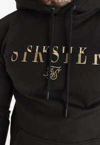 Sudadera negra con el logo bordado en dorado "SIKSILK" y las iniciales, con cordones negros y un bolsillo canguro en la parte delantera.