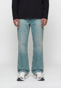 Άτομο που φορά ανοιχτό μπλε τζιν καμπάνα, λευκά και γκρι αθλητικά παπούτσια, και μαύρο μακρυμάνικο τοπ, στέκεται μπροστά σε απλό φόντο.