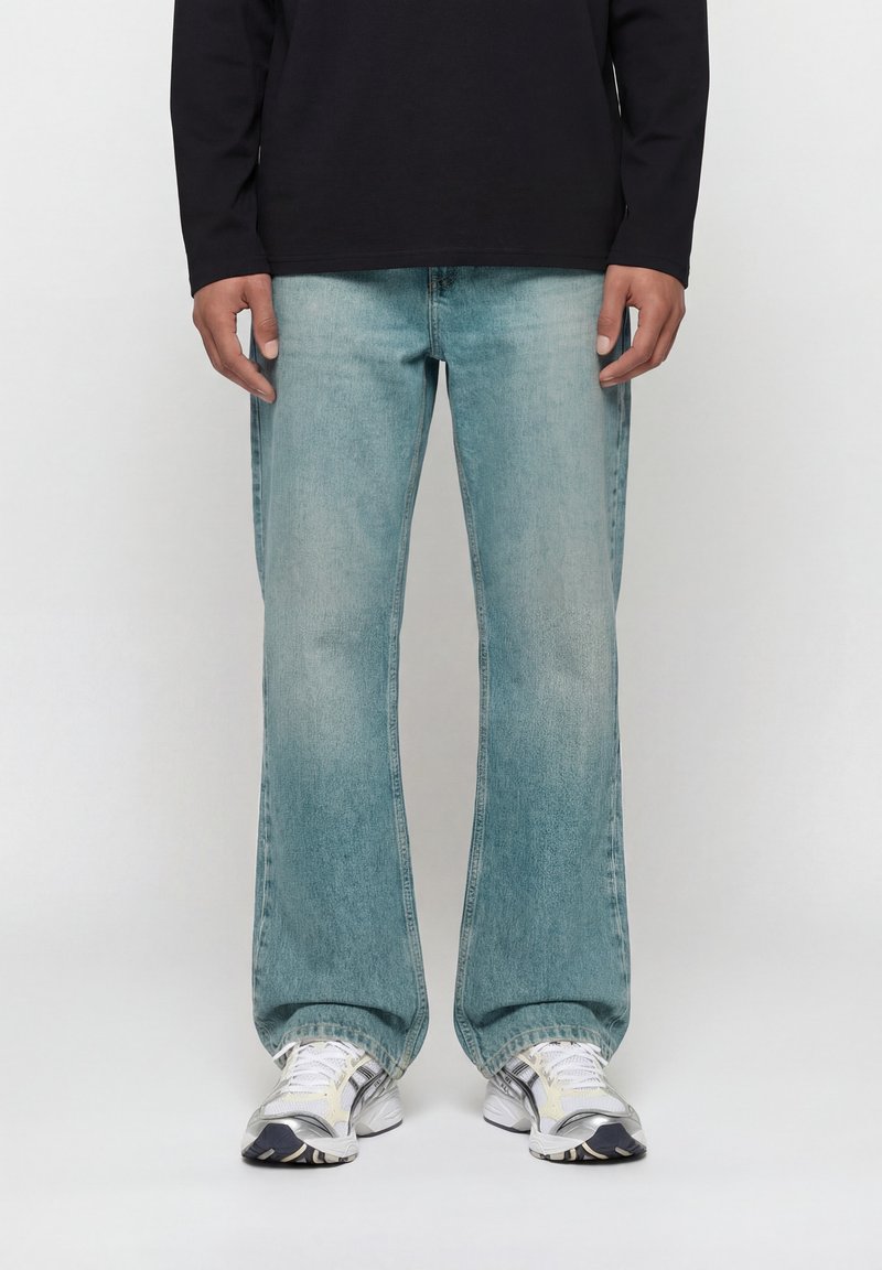 Άτομο που φορά ανοιχτό μπλε τζιν καμπάνα, λευκά και γκρι αθλητικά παπούτσια, και μαύρο μακρυμάνικο τοπ, στέκεται μπροστά σε απλό φόντο.