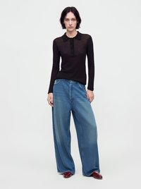 Femme portant un haut noir à manches longues, un jean bleu à jambes larges et des chaussures rouges, debout devant un fond clair uni.