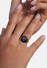 Bague en or avec un visage ovale bleu émaillé, comportant un centre de pierre précieuse triangulaire marron. Texture lisse ; doigts avec un vernis à ongles léger en arrière-plan.