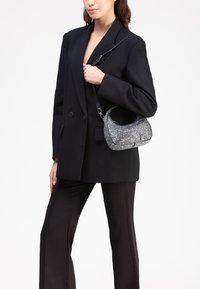 Sac à main noir de type hobo avec un motif de pierres précieuses argentées texturées, une fine bandoulière noire et une forme arrondie, porté sous le bras d'une personne vêtue d'une tenue noire.