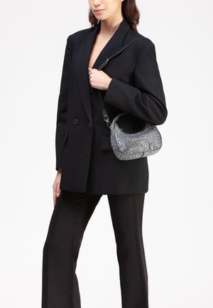 Borsa hobo nera con un motivo testurizzato di gemma argentata, sottile tracolla nera e forma arrotondata, tenuta sotto il braccio di una persona in abbigliamento nero.