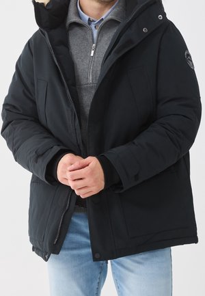 Homme portant une veste matelassée noire par-dessus un pull gris zippé et un jean bleu clair, les mains jointes devant.