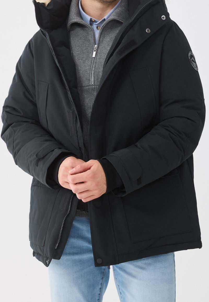 Homme portant une veste matelassée noire par-dessus un pull gris zippé et un jean bleu clair, les mains jointes devant.