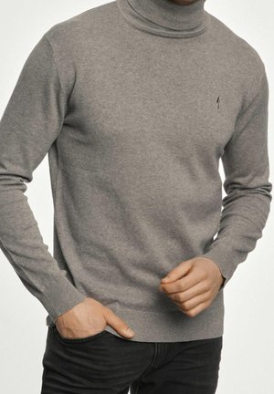 Homme portant un pull gris à col roulé à manches longues et un pantalon noir, une main dans la poche et l'autre légèrement levée.