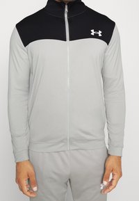 Homme portant une veste de sport zippée gris clair et noir et un pantalon assorti avec des logos Under Armour blancs sur la poitrine et la cuisse.