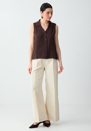 Vrouw met een mouwloze donkerbruine blouse met knopen, crèmekleurige broek met wijde pijpen, donkere hakken en ronde oorbellen, staand tegen een effen achtergrond.