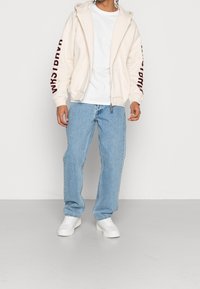 Světle béžová mikina na zip s nápisem "WRSTBHVR" v barvě burgundy na rukávu, doplněná světle modrými džínami a bílými teniskami.