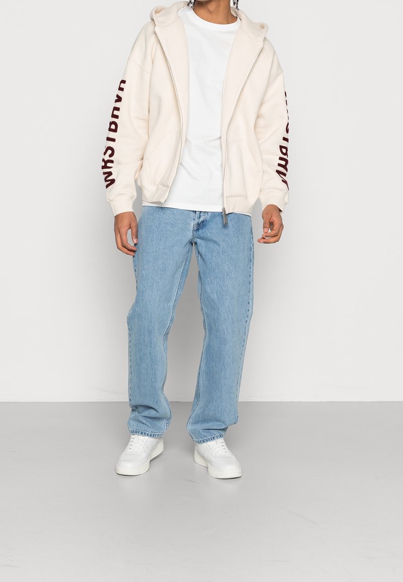Světle béžová mikina na zip s nápisem "WRSTBHVR" v barvě burgundy na rukávu, doplněná světle modrými džínami a bílými teniskami.