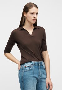 Νεαρή γυναίκα με ίσια καστανά μαλλιά, ντυμένη με καφέ πλεκτό τοπ με κοντά μανίκια και ανοιχτόχρωμο γαλάζιο τζιν ψηλόμεσο, στέκεται με το ένα χέρι στην τσέπη.