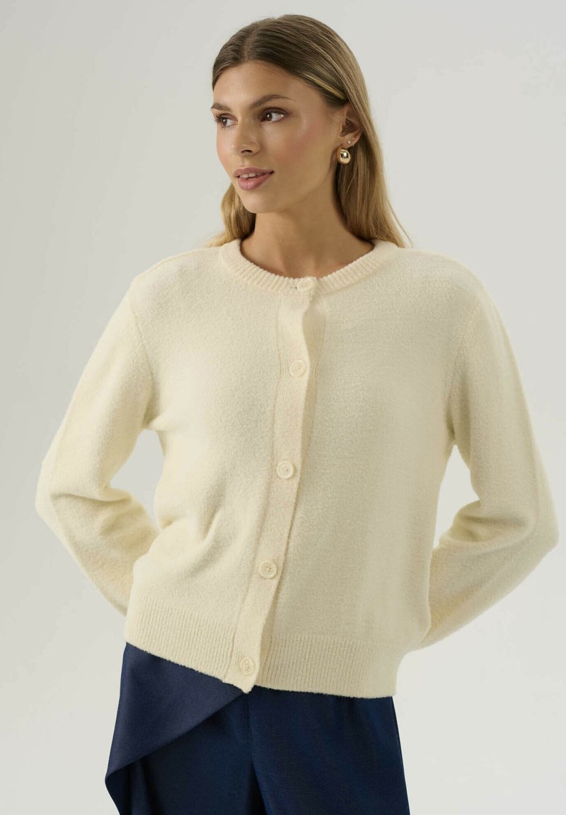 Donna con lunghi capelli biondi che indossa orecchini a cerchio dorati, un cardigan crema abbottonato e pantaloni blu scuro con orlo asimmetrico.