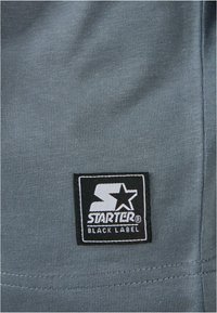 Grå teksturert stoff med et svart "STARTER BLACK LABEL" logo-lapp, med hvite bokstaver og et stjernesymbol.
