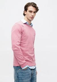 Młody mężczyzna w różowym swetrze na jasnoniebieskiej koszuli i niebieskich dżinsach, stojący z jedną ręką w kieszeni, patrzący w kamerę.