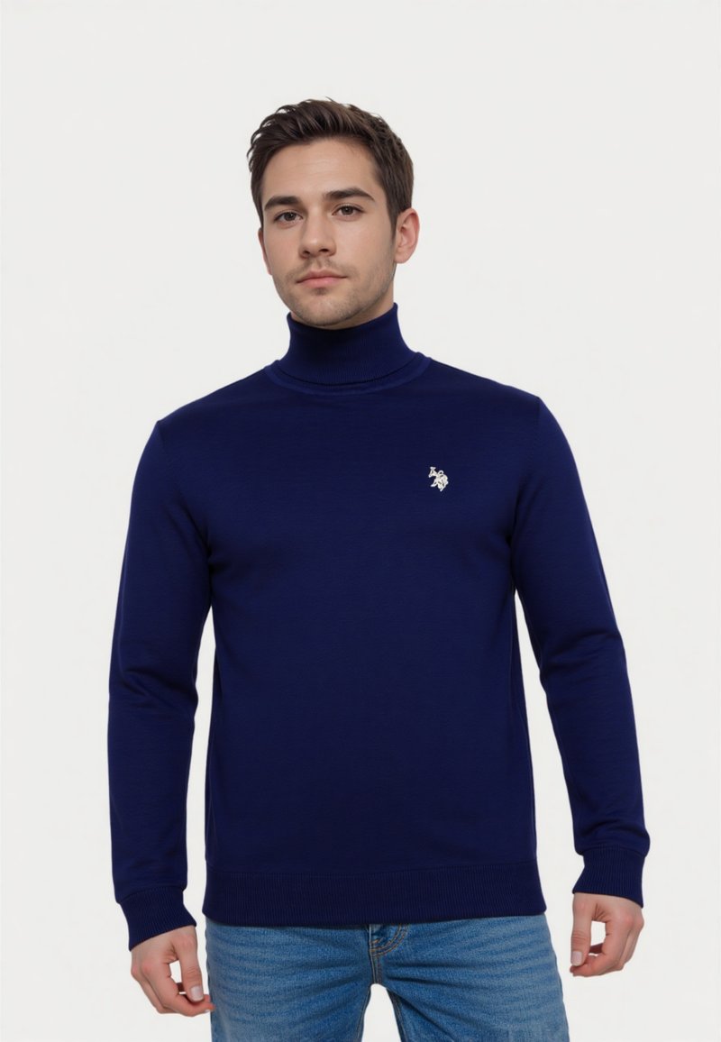 Joven vestido con un suéter de cuello alto azul marino con un pequeño emblema blanco en el pecho, combinado con jeans azules, de pie frente a un fondo blanco.