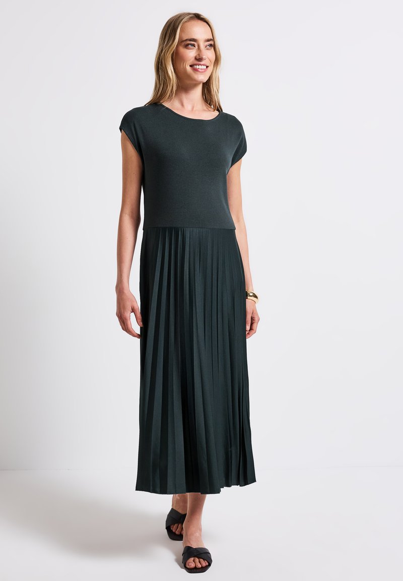 Robe maxi vert foncé avec un corsage ajusté et une jupe plissée. Fabriquée en tissu doux, avec des manches courtes et un col rond.