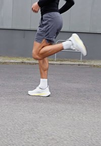 Scarpe da corsa bianche con accenti gialli, pantaloni corti grigi e calze bianche in movimento su una superficie asfaltata, che mostrano gambe muscolose e una postura attiva.