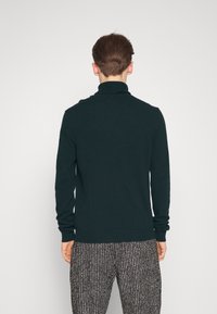 Mörkgrön, långärmad tröja med hög krage, slät textur. Bärs med mönstrade, löst sittande svarta och vita byxor, sedd bakifrån.