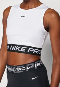 Biały sportowy krótki top o dopasowanym kroju, z czarnym logo Nike i czarną opaską z napisem "NIKE PRO", zestawiony z czarnymi legginsami.