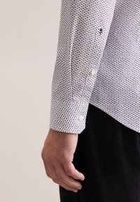 Witte blouse met een klein zwart en grijs patroon, met knoopmanchetten, gestructureerde stof en een zwart accent dicht bij de manchette.