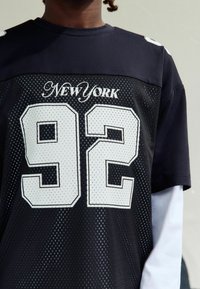 Inimene, kellel on seljas must spordisärk valge võrgu ja tekstiga "New York", suur number 92, valge pika varrukaga särgi peal, lihtsa taustaga.