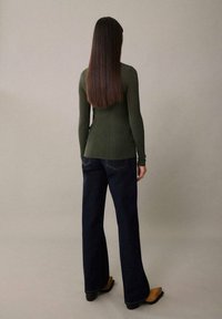 Haut à manches longues vert foncé côtelé, associé à un jean large taille haute en denim foncé et à des bottines à talons couleur tan. Fond neutre.