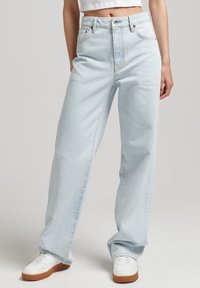 Jeans de tiro alto de color azul claro, con pernera ancha, fabricados en denim, presentan un diseño clásico de cinco bolsillos y sutiles detalles de costura. Combinados con zapatillas blancas.