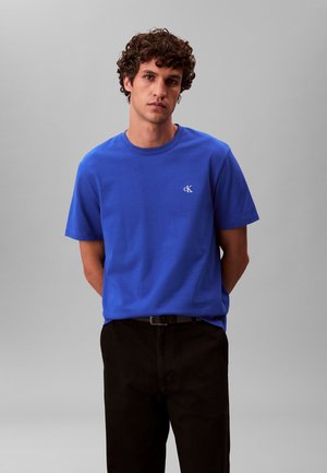 Ung mand med krøllet hår iført en blå T-shirt med korte ærmer og en lille hvid "cK"-logo samt sorte bukser mod en ensfarvet grå baggrund.