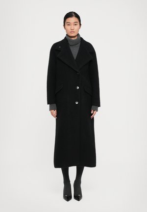 Woman wearing a long black coat with large lapels, buttoned front, over a grey turtleneck sweater, dark tights, and black pointed heels.