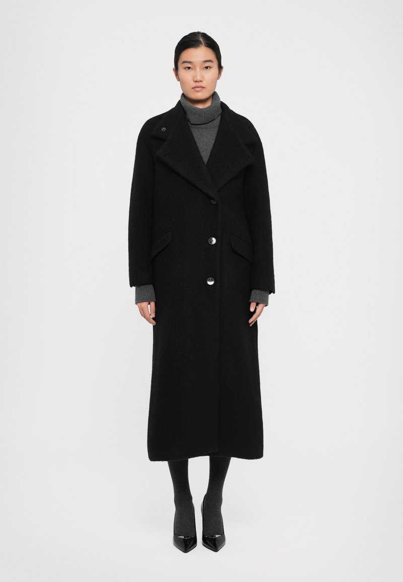 Femme portant un long manteau noir avec de grands revers, boutonné à l'avant, sur un pull à col roulé gris, des collants sombres et des escarpins noirs à bout pointu.