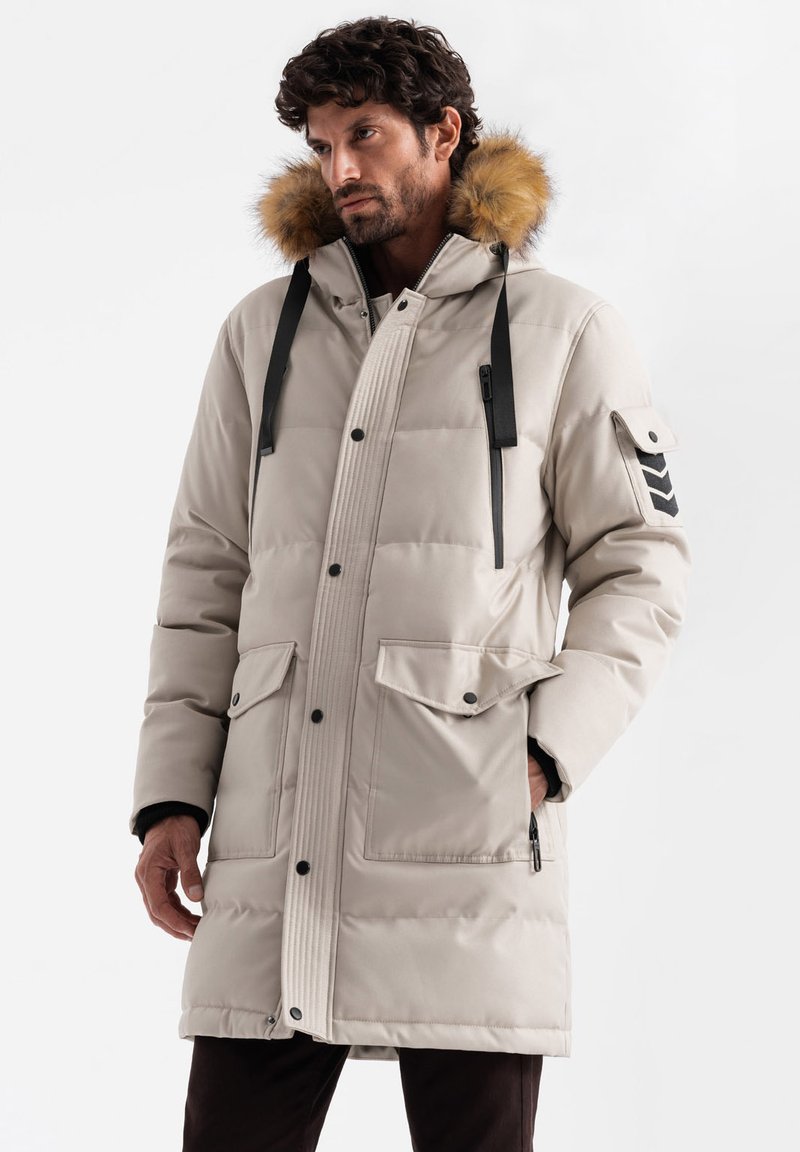 Parka imbottita beige chiaro con cappuccio foderato in pelliccia, doppie tasche frontali, dettagli con cerniere e strisce nere a contrasto sulle spalle.