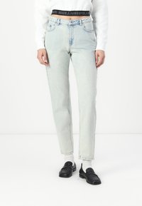 Jeans de cintura alta de cor azul clara com um corte reto, apresentando bolsos frontais e um efeito desbotado. Usados com um suéter branco e mocassins pretos.