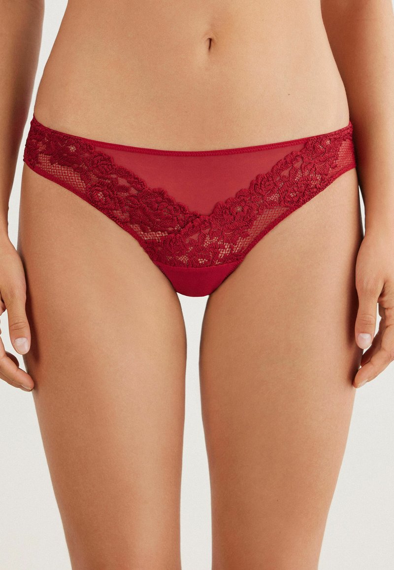 Bikini taille basse en dentelle rouge et tissu transparent, avec des détails en dentelle florale le long de la ceinture et des côtés. Texture douce et extensible pour un confort optimal.