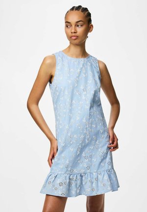 Jeune femme aux cheveux tressés portant une robe sans manches bleu clair avec des motifs floraux découpés blancs et un ourlet à volants.