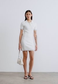 Λευκό φόρεμα πόλο με υφασμάτινο μοτίβο, αντίθετο τελείωμα, κοντά μανίκια και εφαρμοστή σιλουέτα. Αξεσουάρ: μικρή λευκή τσάντα.