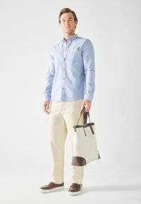 Light blue button-up shirt with a chest pocket, cream-colored pants, and brown shoes. Holds a cream tote bag with brown accents.