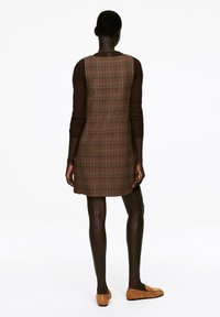 Braun karierter Kleid mit taillierter Form, ärmellosem Design und einer Rückennaht. Das Modell trägt ein braunes Langarmoberteil und passende Slipper.