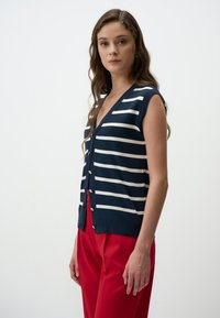 Marineblauwe mouwloze cardigan met horizontale witte strepen, v-hals en knopenfront. Gecombineerd met rode broek van een gestructureerde stof.